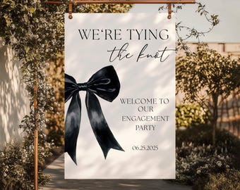 May include: A white sign with the text "WE'RE TYING the knot" and "WELCOME TO OUR ENGAGEMENT PARTY 06.25.2025". A large black bow illustration is centered on the sign. The sign is hanging from a copper-colored rod.