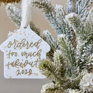 May include: White house-shaped Christmas ornament with gold lettering that reads "ordered too much takeout 2020". The ornament has gold paint splatters on it and is hanging from a white ribbon on a snowy artificial Christmas tree.