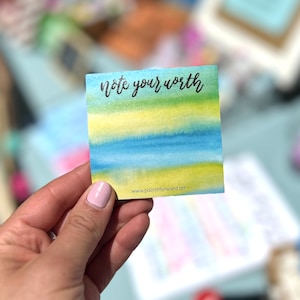 May include: A watercolor note card with a blue, yellow, and green ombre design. The card has the text "note your worth" in black lettering and the website "www.paint itforward.art" printed at the bottom.
