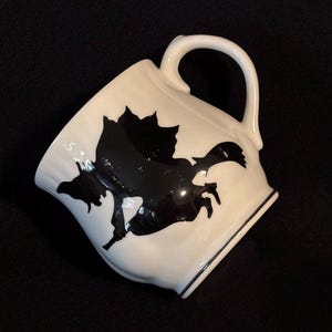 May include: White ceramic mug with a black silhouette of a witch riding a broom. The mug has a curved handle and a black trim around the rim and base. The design is a Halloween or fantasy theme.