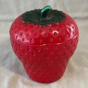 May include: A red ceramic strawberry-shaped container with a green stem and leaf design. The container has a lid and is covered in small, textured bumps. The strawberry-themed item is likely a decorative storage piece.