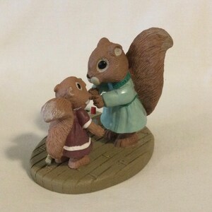 Puede incluir: Una figurita de ardilla marr&oacute;n y beige con un vestido azul y verde est&aacute; sosteniendo la mano de una figurita de ardilla marr&oacute;n y beige m&aacute;s peque&ntilde;a con un vestido granate. Las figuritas est&aacute;n de pie sobre una base de madera marr&oacute;n.