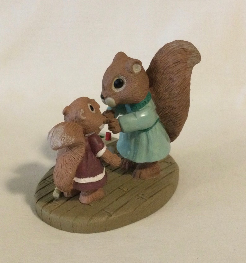 Puede incluir: Una figurita de ardilla marr&oacute;n y beige con un vestido azul y verde est&aacute; sosteniendo la mano de una figurita de ardilla marr&oacute;n y beige m&aacute;s peque&ntilde;a con un vestido granate. Las figuritas est&aacute;n de pie sobre una base de madera marr&oacute;n.