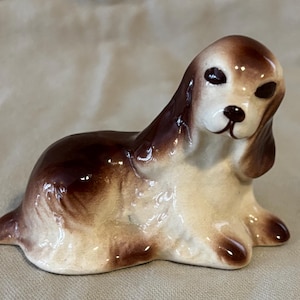 May include: A ceramic figurine of a dog in a seated position. The dog has brown and white coloring, with brown ears and a brown patch on its back. The figurine has a glossy finish and is likely a decorative item.