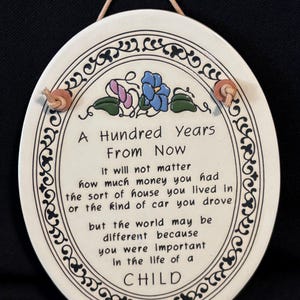 May include: An oval ceramic wall plaque with a cream-colored background and a black decorative border. It features a hand-painted blue and purple floral design at the top and black text that reads: A Hundred Years From Now it will not matter how much money you had the sort of house you lived in or the kind of car you drove but the world may be different because you were important in the life of a CHILD. It includes a tan rope hanger.