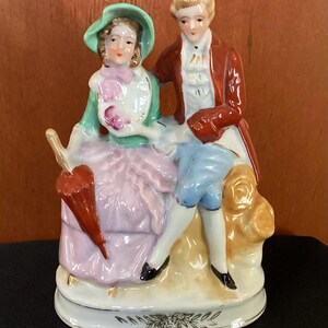 May include: A porcelain figurine depicting a seated couple in 18th-century attire. The woman wears a pink dress and green hat, holding a red umbrella. The man is in a red coat and blue breeches. The base is white with gold accents.