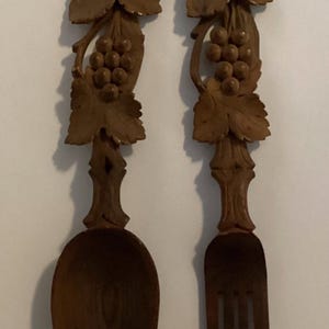 May include: A set of two wooden serving utensils, a spoon and a fork, with intricate carvings of grapevines and grapes. The utensils are brown and have a rustic appearance.