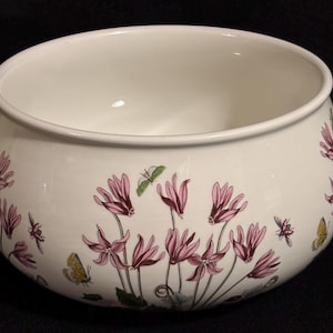 May include: A white ceramic bowl with a floral design featuring pink flowers, green insects, and yellow butterflies. The bowl has a rounded shape and a smooth, glossy finish. The floral pattern is repeated around the exterior.