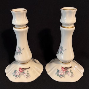 May include: Pair of off-white ceramic candlesticks with gold trim. Each candlestick features a bird and floral design in pink, green, and red. The base is scalloped, and the overall design is elegant and classic.