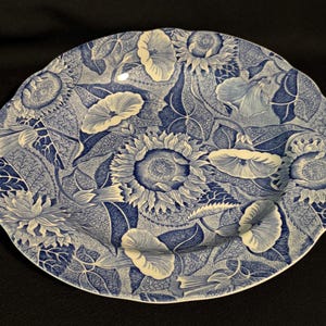 May include: A round ceramic plate with a scalloped edge featuring a detailed blue and white transferware pattern of sunflowers and morning glory flowers. The intricate botanical design covers the entire surface with dense foliage and petals.