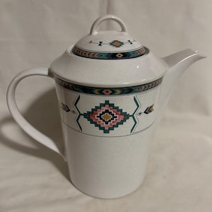 May include: A white ceramic teapot with a lid and handle. The teapot features a speckled finish and is adorned with a geometric pattern in teal, pink, yellow, and black. The spout is on the right side.