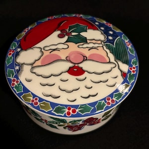 May include: A round, ceramic Christmas box featuring a smiling Santa Claus face on the lid. The box has a blue border with holly and berry accents. Santa's hat is red, and his face is pink and white.