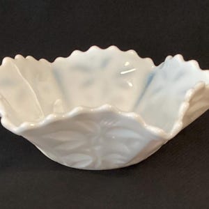 Puede incluir: Un cuenco blanco con borde festoneado y diseño floral. El cuenco tiene forma plegada de pétalo y está hecho de un material brillante y opaco. El cuenco está sobre un fondo oscuro.