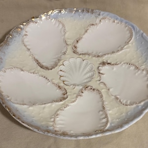 May include: A white ceramic oyster serving platter with six shell-shaped compartments. The platter has a scalloped edge with gold trim and a central seashell design. The rim has a light blue and gold border.
