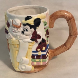 May include: A white ceramic mug with a brown handle. The mug features a 3D design of Mickey Mouse and Santa Claus. The design is painted in various colors, including red, yellow, blue, and green.