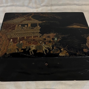 May include: Black rectangular box with a detailed Asian landscape scene on the lid. The scene depicts figures, horses, and a pagoda in shades of gold, brown, and red against a black background. The box has a small, round, red clasp.