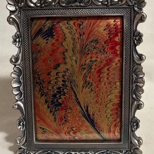 May include: A silver-toned picture frame with an ornate design. The frame has a rectangular opening with a colorful marbled paper inside.