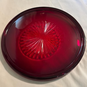May include: A round, ruby red glass plate with a raised rim. The center features a starburst design. The plate is likely vintage and adds a pop of color to any table setting.