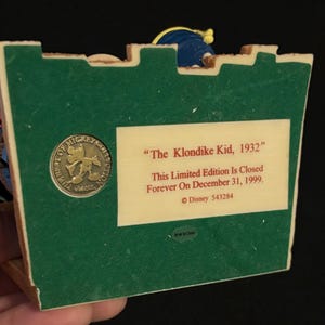 May include: A collectible figurine base with a green surface and a beige rectangular label. The label reads "The Klondike Kid, 1932" and "This Limited Edition Is Closed Forever On December 31, 1999." A circular emblem with Mickey Mouse is also visible.