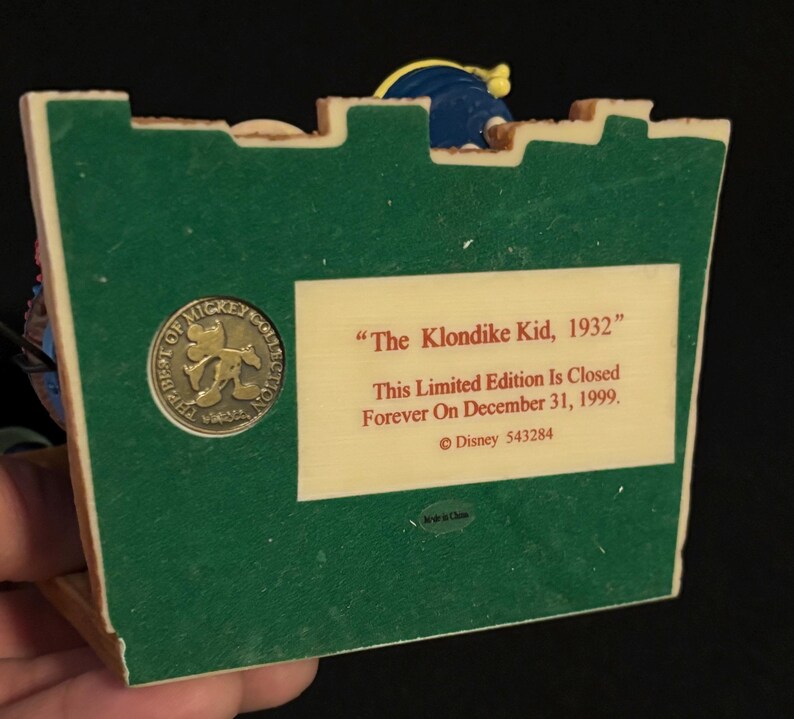 May include: A collectible figurine base with a green surface and a beige rectangular label. The label reads "The Klondike Kid, 1932" and "This Limited Edition Is Closed Forever On December 31, 1999." A circular emblem with Mickey Mouse is also visible.