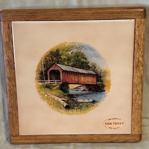 May include: Oak trivet with a framed wooden border. The trivet features a circular image of a covered bridge over a river, surrounded by trees. The Daniels Co. Oak Trivet is printed on the trivet.