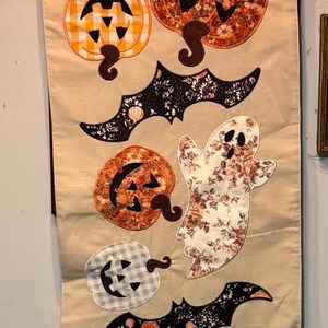 May include: A beige table runner featuring appliqued Halloween motifs including ghosts, pumpkins, and bats. The fabric elements display various patterns such as floral prints and gingham checks. The top label reads SHABBY CHIC.