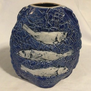 May include: A blue ceramic vase with a textured surface and three white fish designs. The vase is shaped like a rounded rectangle.