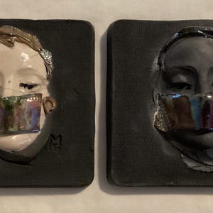 May include: Two ceramic wall art pieces. One features a light-skinned face with a colorful, iridescent mouth covering. The other is a dark-skinned face with similar iridescent mouth covering. Both are set on a dark square background.