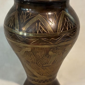 May include: A bronze-colored vase with intricate etched designs. The vase features geometric and stylized figures in a lighter gold color. The vase has a wide body and a narrow base, with a flared rim. A decorative piece for home decor.