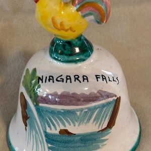 May include: A ceramic bell with a yellow rooster on top. The bell is white with a painted scene of Niagara Falls and the words "NIAGARA FALLS". The rooster has red, orange, and blue tail feathers.