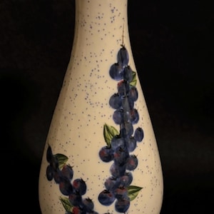 May include: A ceramic vase with a cream-colored base featuring a blue speckled pattern. The vase is decorated with hand-painted clusters of dark blue berries and green leaves. It has a flared rim and a circular base, both finished with a blue sponge-painted effect.