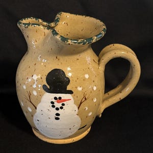 May include: A ceramic pitcher with a speckled tan glaze and a dark green rim. The front features a hand-painted white snowman wearing a black top hat, with a bright orange carrot nose, black button eyes, and twig-like arms amidst white dots.