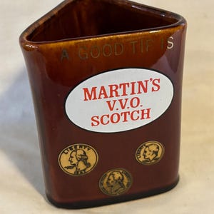 May include: A brown ceramic triangular shaped cup with the words "A Good Time Is" and "Martin's V.V.O. Scotch" printed on it. The cup has four gold coins with the word "Liberty" printed on them.