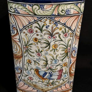 May include: A triangular ceramic vase featuring a white base with hand-painted floral motifs, scrolling foliage, and a central bird illustration in shades of blue, green, yellow, and orange, finished with a dark blue rim at the top edge.