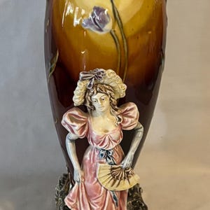 May include: A decorative ceramic vase with a woman figurine. The vase has a brown and yellow background with floral accents. The woman is wearing a pink dress and holding a fan. The vase has two handles.