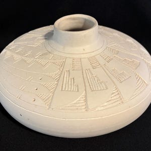 May include: A low-profile, matte white ceramic vessel with a wide, rounded body and a central circular opening. The surface features intricate, carved geometric patterns including stepped motifs, linear hatching, and stylized fish shapes.