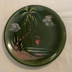 May include: A round, dark green decorative plate with a tropical scene. The plate features a palm tree, a pink flamingo, and stylized clouds. The words "Miami Beach Fla" are written on the plate.