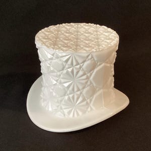 May include: A white, decorative top hat-shaped container. The hat features a textured, geometric pattern on the crown and sides, with a smooth, wide brim. The container is made of a glossy, opaque material.