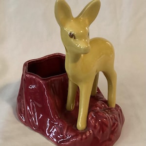 May include: A ceramic planter featuring a yellow deer figurine standing on a burgundy base. The deer has large ears and a slender body. The base is a deep burgundy color with a textured appearance, and has a small opening for planting.