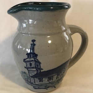 May include: A gray ceramic pitcher with a blue and white church scene painted on the side. The pitcher has a green glazed rim.