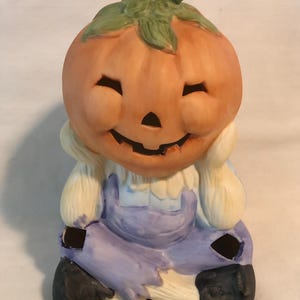 May include: A ceramic pumpkin figurine with a smiling face, wearing blue overalls and sitting with its legs crossed. The pumpkin has a green stem and a carved out face with a candle holder.