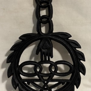 May include: Black cast iron trivet with a decorative design. The trivet features a hand, a series of interlocking circles, and a leafy border. It has a chain for hanging. A functional and decorative kitchen item.