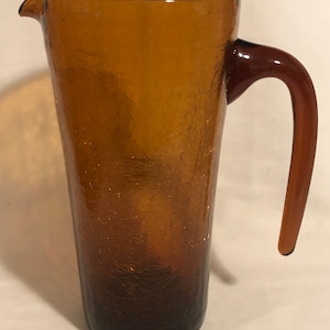 May include: A brown glass pitcher with a textured surface and a brown handle. The pitcher is tall and cylindrical.