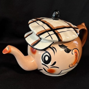 May include: A novelty teapot with a cartoon face design. The teapot is tan with black, white, and brown accents. The lid is a hat with a black knob. The teapot has a handle and a spout.