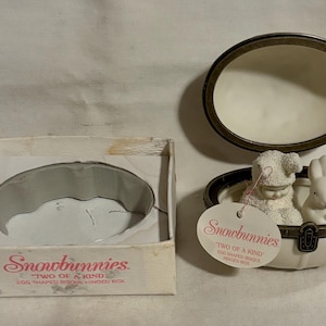 May include: A white ceramic egg-shaped box with a hinged lid. The box is decorated with gold trim and features two small snowmen figurines inside. The box is labeled "Snowbunnies Two of a Kind Egg Shaped Special Hinged Box".