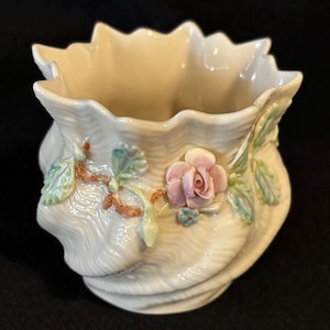 May include: Cream-colored ceramic vase with a ruffled rim and a textured, swirling design. The vase is adorned with a pink rose, green and blue leaves, and small blue flowers. The vase is a decorative item.