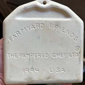 Puede incluir: Placa rectangular de cer&aacute;mica blanquecina con la inscripci&oacute;n "FARM YARD FRIENDS" en arco. Tambi&eacute;n se lee "THE PAMPERED CHEF LTD.", "1994" y "USA". Tiene un agujero en la parte superior para colgar.