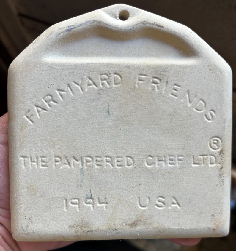 Puede incluir: Placa rectangular de cer&aacute;mica blanquecina con la inscripci&oacute;n "FARM YARD FRIENDS" en arco. Tambi&eacute;n se lee "THE PAMPERED CHEF LTD.", "1994" y "USA". Tiene un agujero en la parte superior para colgar.
