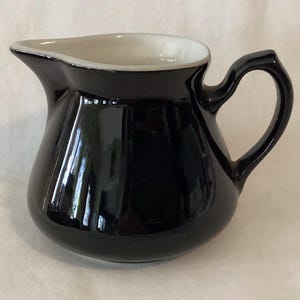 May include: A glossy black ceramic creamer pitcher with a white interior and a curved handle. The pitcher has a rounded body and a spout for easy pouring. The handle is arched and attached to the side of the pitcher.
