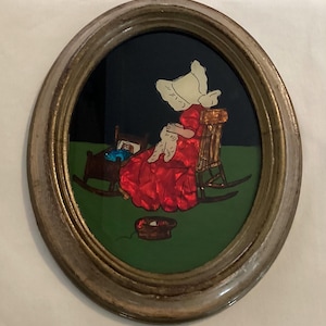 May include: Oval framed artwork depicting a woman in a red dress and white bonnet, seated in a rocking chair. A cradle with a blue blanket and a basket of yarn are also in the scene. The background is green and black.